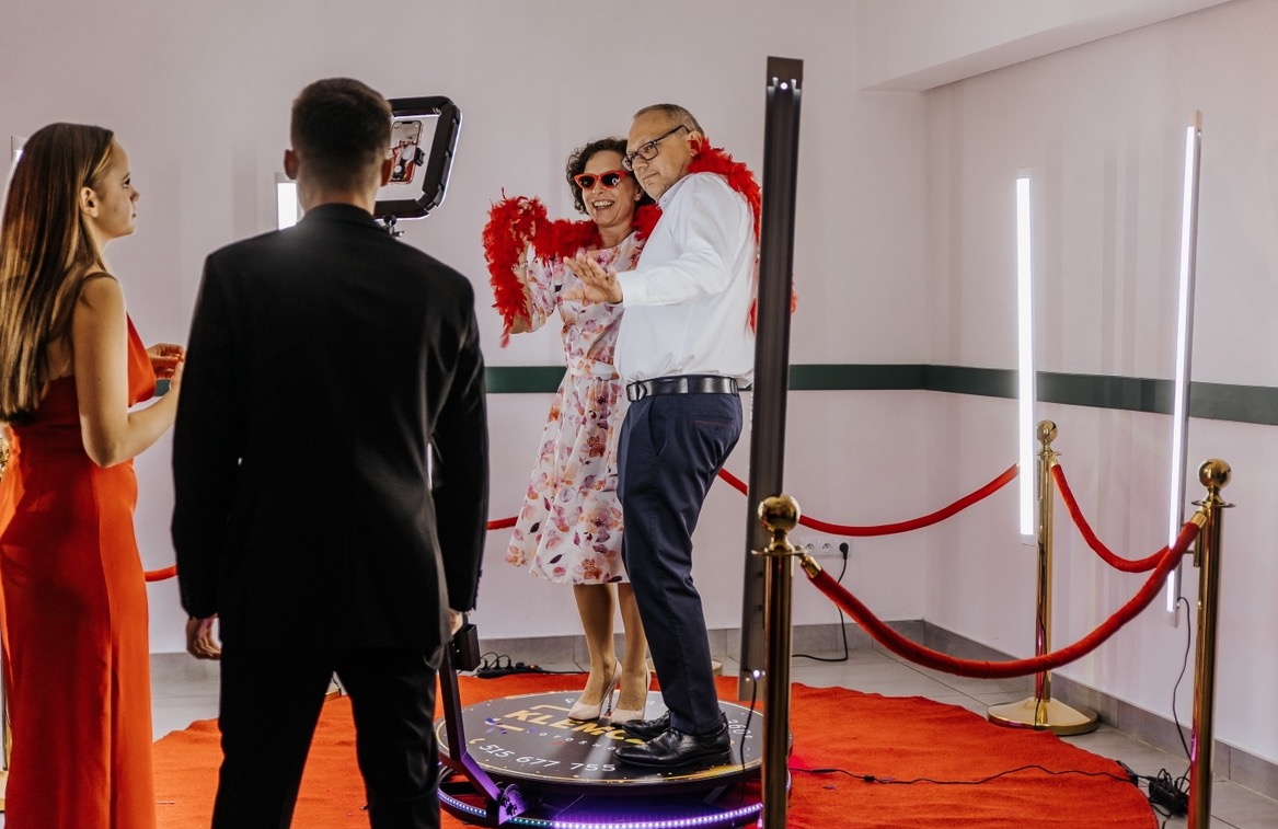 Para w czerwonych boa i okularach tańczy na obrotowej platformie fotobudki 360, otoczonej czerwonym dywanem i złotymi słupkami, podczas gdy mężczyzna w garniturze obsługuje urządzenie, a kobieta...