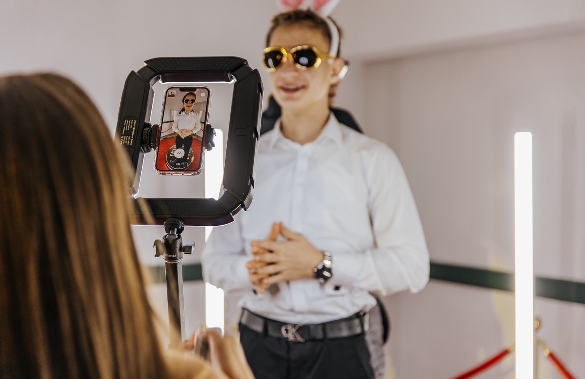 Osoba z króliczymi uszami i złotymi okularami pozuje przed smartfonem umieszczonym w ringu świetlnym, widocznym z perspektywy osoby obsługującej urządzenie.