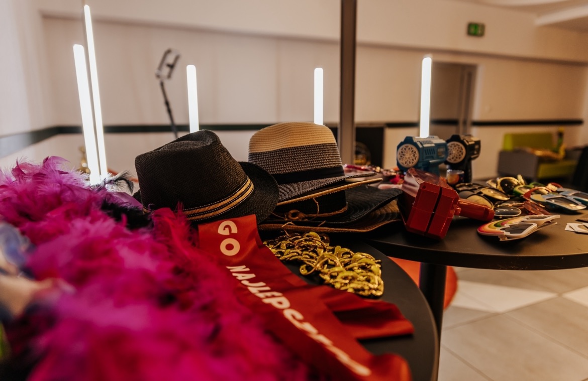 Rekwizyty na dwóch okrągłych stolikach przygotowane do sesji zdjęciowej: kolorowe boa z piór, kapelusze, złote łańcuchy, śmieszne okulary, czerwona szarfa z napisem oraz zabawki.