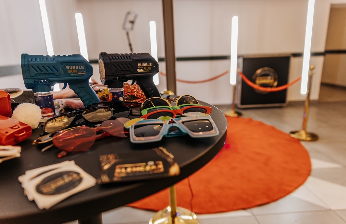 Akcesoria do sesji zdjęciowej: pistolety na bańki mydlane, okulary w różnych kształtach i rekwizyty leżące na okrągłym stole, w tle widoczny automat do zdjęć z czerwonym sznurem.