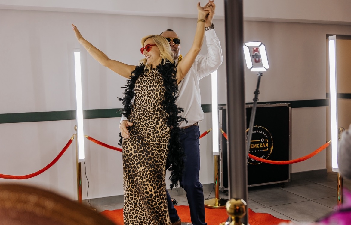 Uśmiechnięta kobieta w panterkowej sukience i boa z piór, pozująca z mężczyzną w okularach w kształcie serc, przed tłem z czerwonym dywanem i lampami, prawdopodobnie podczas imprezy...