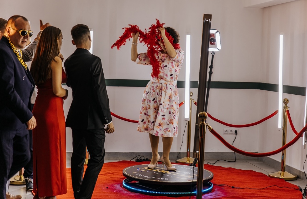 Kobieta w kwiecistej sukience i z czerwonym boa pozuje na obrotowej platformie w otoczeniu gości i czerwonego dywanu, oświetlona lampami LED