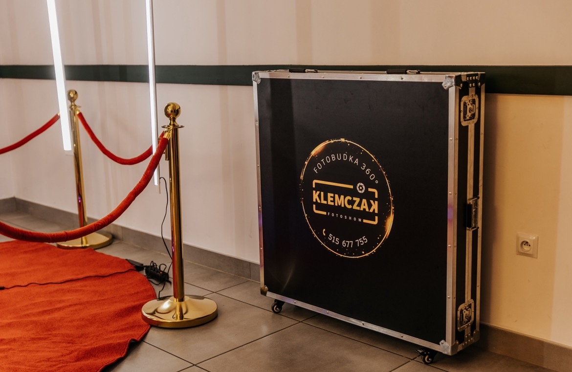 Czarna skrzynia transportowa z logo firmy oferującej fotobudkę 360 stopni, ustawiona obok złotych słupków z czerwonym sznurem i czerwonym dywanem.