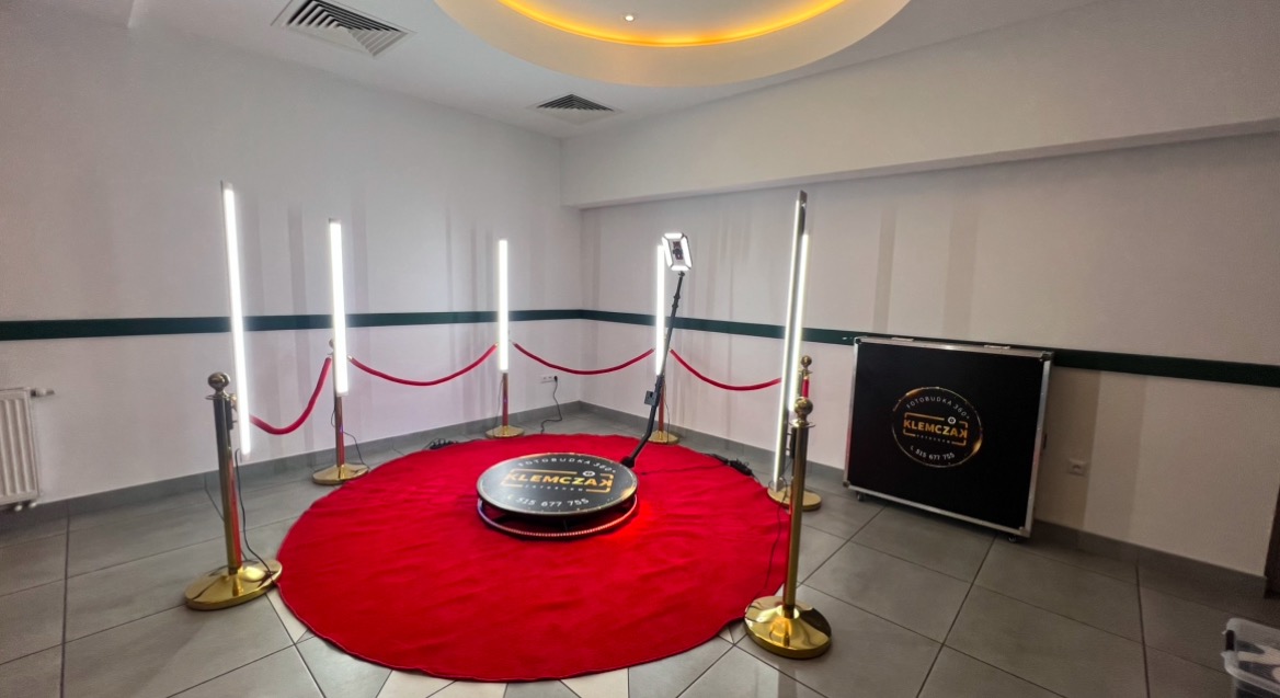 Rozstawiona fotobudka 360 na czerwonym dywanie, otoczona złotymi słupkami z czerwonym sznurem, oświetlona czterema lampami LED w białym pomieszczeniu z podświetlanym sufitem.