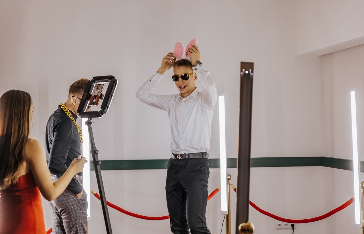Uczestnicy imprezy pozują z rekwizytami, uszy królika, przed obiektywem w stanowisku do zdjęć z lampami LED, na tle białej ściany z czerwonym sznurem.