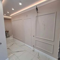 Michał Majewski - Wykończone wnętrze korytarza z białymi płytkami imitującymi marmur, kremowymi ścianami z ozdobnymi listwami i oświetleniem LED pod sufitem, widoczny worek z materiałami budowlanymi w tle.