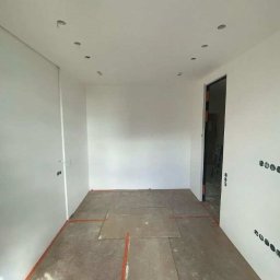 Michał Majewski - Pokój w trakcie remontu: białe ściany z widocznymi otworami na oświetlenie w suficie, zabezpieczona podłoga, przygotowania do malowania, widoczne gniazdka elektryczne.