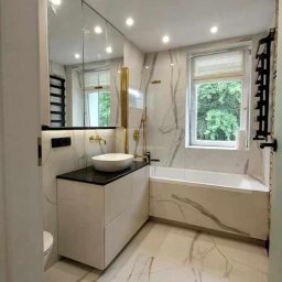 Michał Majewski - Nowoczesna łazienka z marmurowymi płytkami, białą umywalką nablatową na szafce z czarnym blatem, złotą armaturą i grzejnikiem drabinkowym.