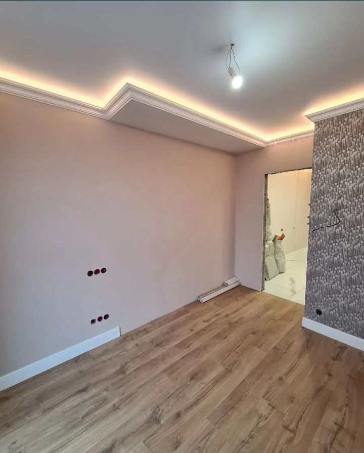 Wykończone wnętrze pokoju w Grudziądzu z panelami na podłodze, pomalowanymi ścianami w odcieniach różu, ozdobnym sufitem z oświetleniem LED i fragmentem ściany z tapetą. Widoczne puste otwory...