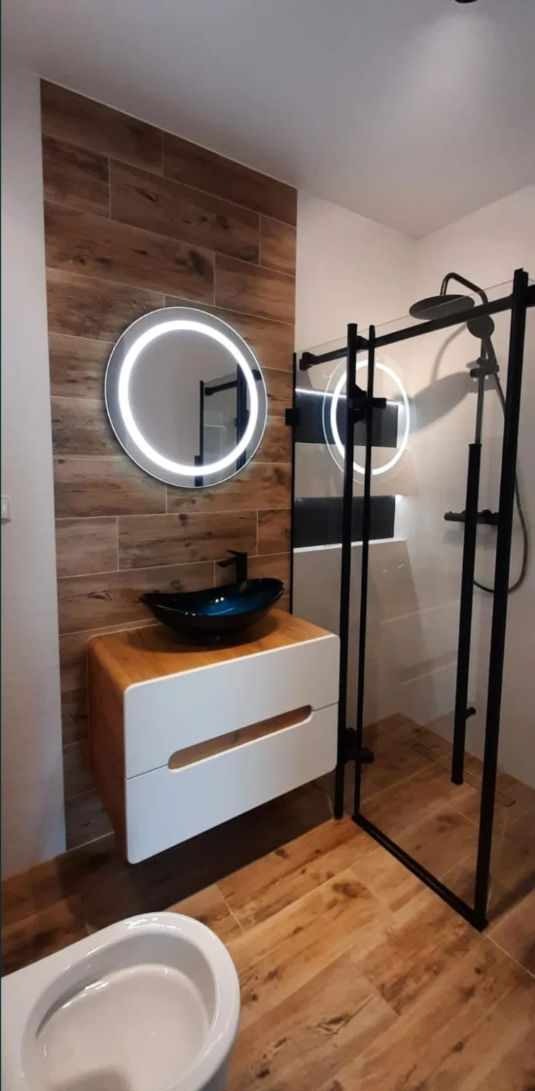Nowoczesna łazienka z drewnopodobnymi płytkami, okrągłym lustrem z podświetleniem LED, czarną szklaną kabiną prysznicową i umywalką w kształcie misy na drewnianej szafce z białymi szufladami.