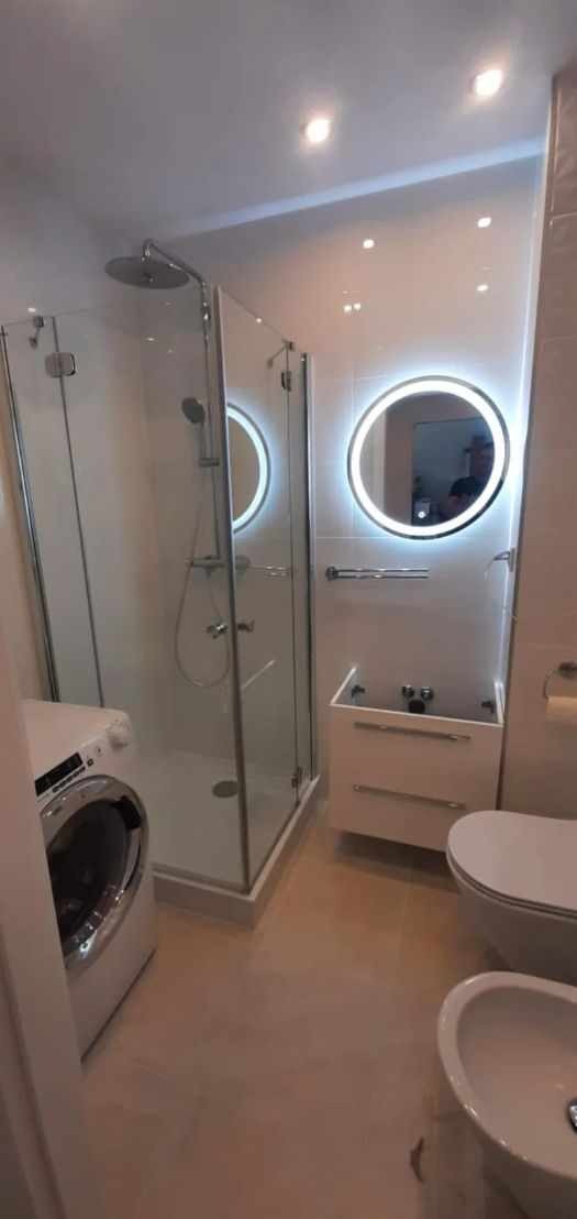 Nowoczesna łazienka z kabiną prysznicową, okrągłym lustrem podświetlanym LED, białą umywalką z szafką, toaletą i bidetem, oraz pralką w rogu.