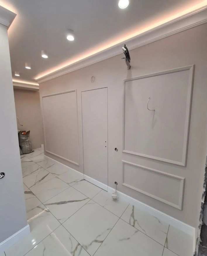 Wykończone wnętrze korytarza z białymi płytkami imitującymi marmur, kremowymi ścianami z ozdobnymi listwami i oświetleniem LED pod sufitem, widoczny worek z materiałami budowlanymi w tle.