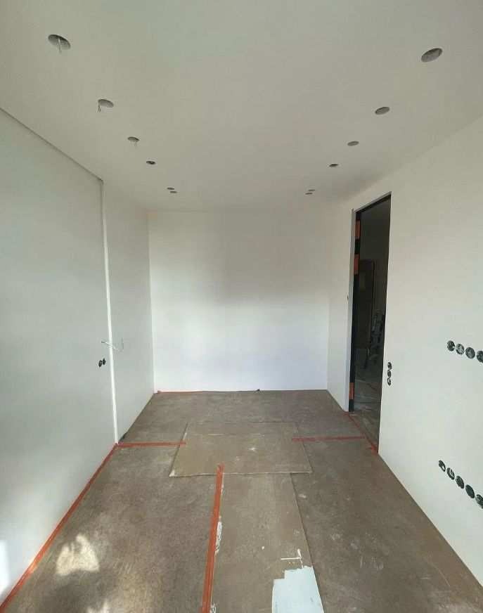 Pokój w trakcie remontu: białe ściany z widocznymi otworami na oświetlenie w suficie, zabezpieczona podłoga, przygotowania do malowania, widoczne gniazdka elektryczne.