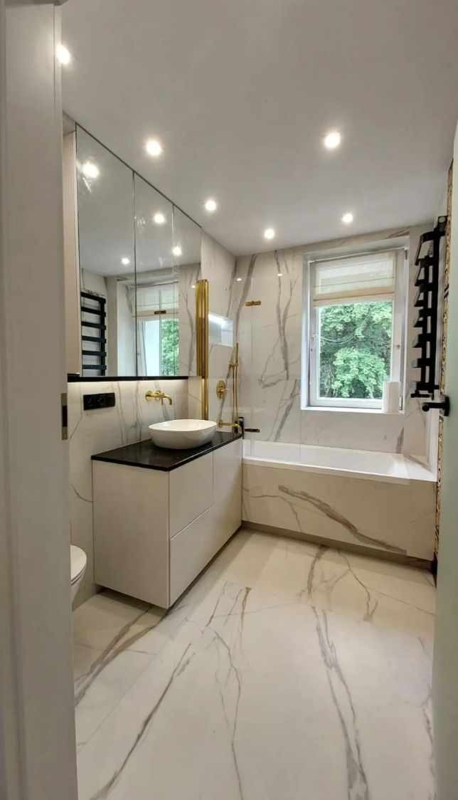 Nowoczesna łazienka z marmurowymi płytkami, białą umywalką nablatową na szafce z czarnym blatem, złotą armaturą i grzejnikiem drabinkowym.