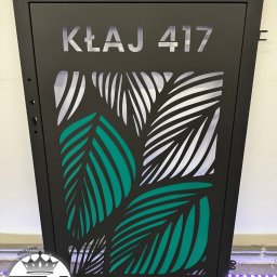 KM Design DOMINIKA KORYCIŃSKA - Czarna metalowa skrzynka na listy z wycinanym laserowo wzorem liści palmowych i numerem domu 'KŁAJ 417', oświetlona od wewnątrz.