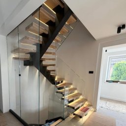 KM Design DOMINIKA KORYCIŃSKA - Nowoczesne schody drewniane z podświetleniem LED i szklaną balustradą, widok z dołu, minimalistyczny design, jasne wnętrze.