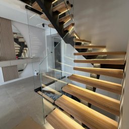 KM Design DOMINIKA KORYCIŃSKA - Nowoczesne schody drewniane z podświetleniem LED i szklaną balustradą, widok z dołu, eleganckie wykończenie wnętrza, minimalistyczny design.