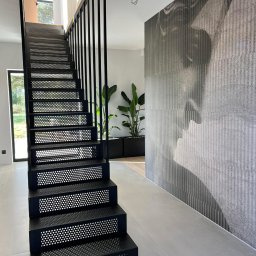 KM Design DOMINIKA KORYCIŃSKA - Nowoczesne schody metalowe w kolorze czarnym z perforowanymi stopniami, minimalistyczna balustrada, w tle ściana z abstrakcyjnym wzorem i rośliny doniczkowe.