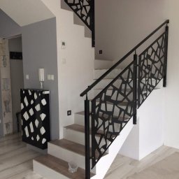 KM Design DOMINIKA KORYCIŃSKA - Nowoczesne schody z czarną, metalową balustradą o geometrycznym wzorze w jasnym wnętrzu. Widoczne płytki imitujące drewno na stopniach i podłodze.