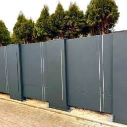 KM Design DOMINIKA KORYCIŃSKA - Nowoczesna, przesuwna brama wjazdowa w kolorze grafitowym z pionowymi, białymi zdobieniami, na tle zielonych tuj i kostki brukowej. Widoczne mocowania bramy.