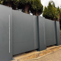 KM Design DOMINIKA KORYCIŃSKA - Nowoczesna, przesuwna brama wjazdowa w kolorze grafitowym z minimalistycznymi, pionowymi zdobieniami, widoczna z boku, na tle zielonych drzew i kostki brukowej.