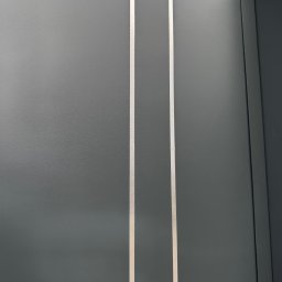 KM Design DOMINIKA KORYCIŃSKA - Fragment nowoczesnego, grafitowego ogrodzenia metalowego z dwoma pionowymi, jasnymi paskami. Widoczny narożnik ogrodzenia i fragment roślinności w tle.