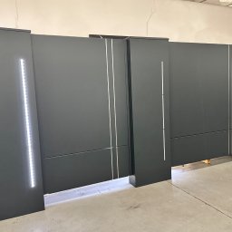 KM Design DOMINIKA KORYCIŃSKA - Nowoczesna, grafitowa brama wjazdowa z subtelnym oświetleniem LED, widoczna w hali produkcyjnej. Minimalistyczny design z pionowymi akcentami.