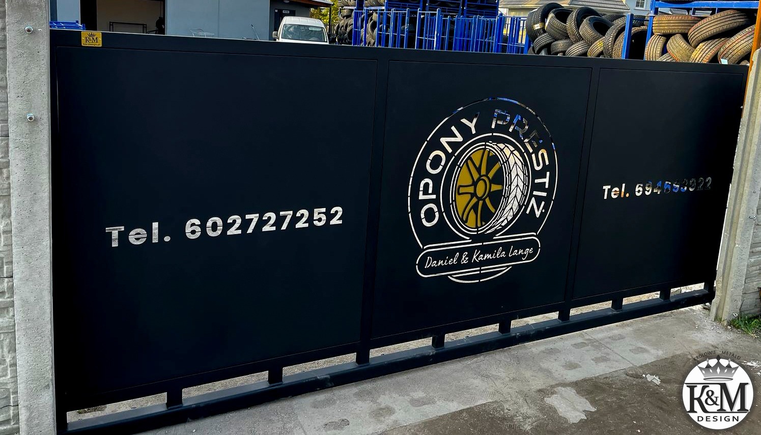 Czarna brama wjazdowa z logo firmy oponiarskiej i numerami telefonów, widoczne opony w tle.