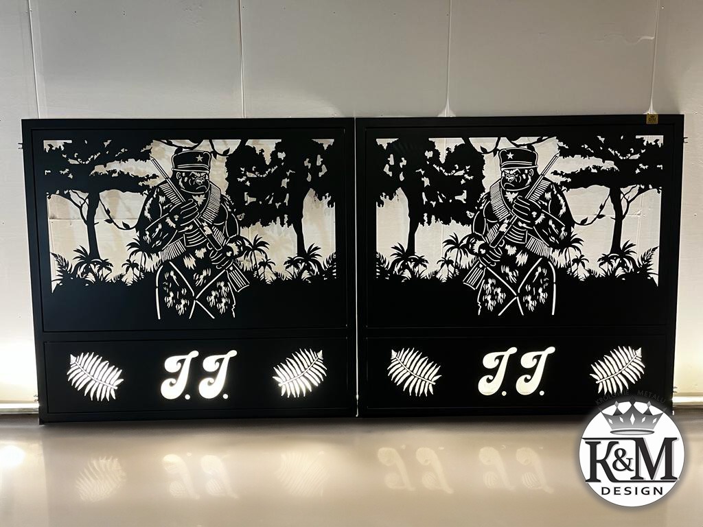 Dwie czarne metalowe bramy wjazdowe z wycinanym laserowo wzorem żołnierza w dżungli, podświetlane od tyłu, z inicjałami 'JJ' i motywem liści paproci, widoczne odbicie na podłodze.