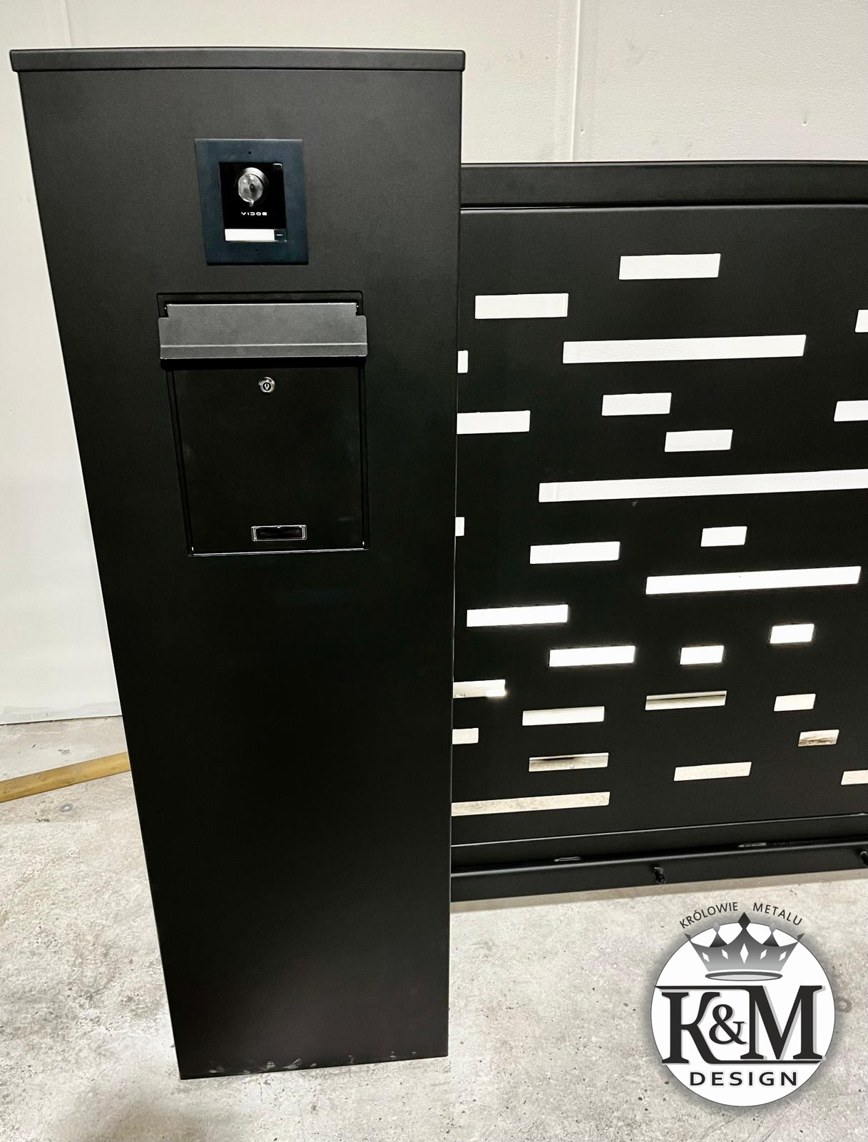 Czarny, nowoczesny słupek z wideodomofonem i skrzynką na listy, przylegający do ażurowego panelu ogrodzeniowego z poziomymi, prostokątnymi prześwitami.