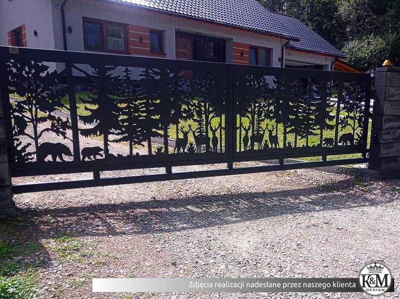 Czarna brama wjazdowa z motywem wycinanego lasu, przedstawiająca sylwetki drzew, jeleni i niedźwiedzi, zamontowana przed domem z szarą dachówką.
