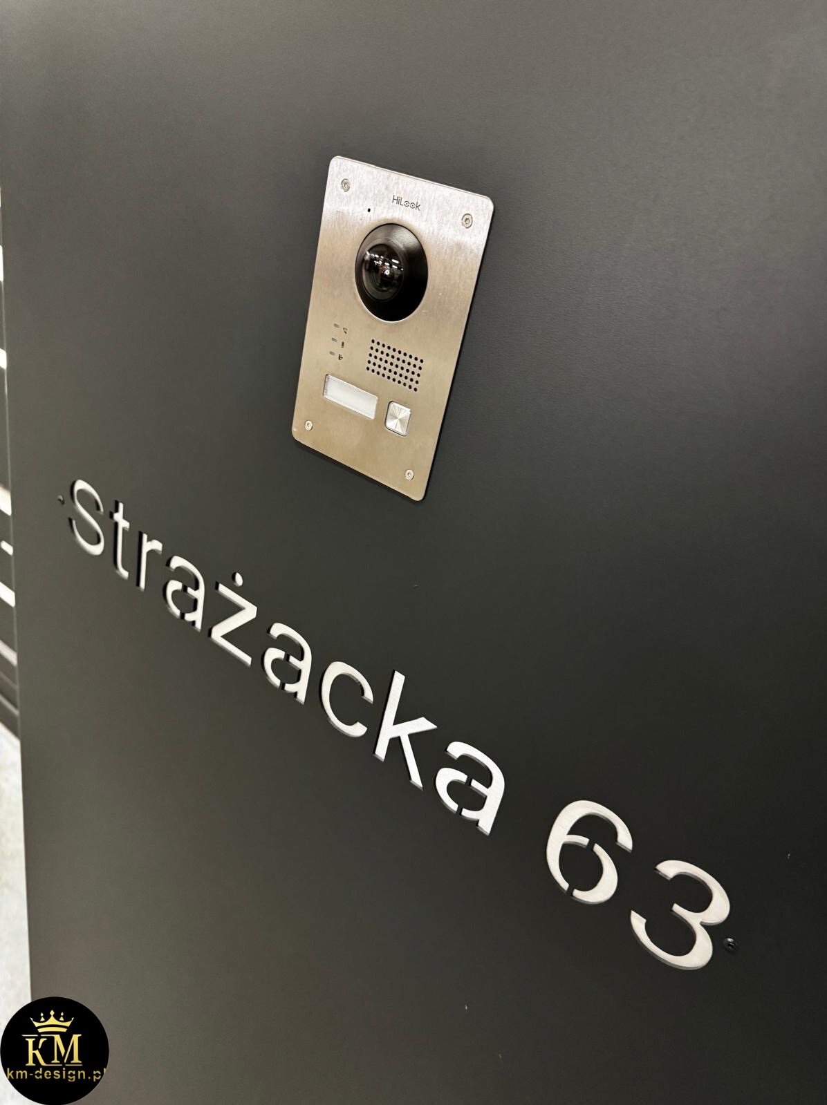 Czarny panel z adresem 'Strażacka 63' i srebrnym domofonem HiLook. Minimalistyczny design, nowoczesne wykończenie, oświetlenie podkreśla detale.