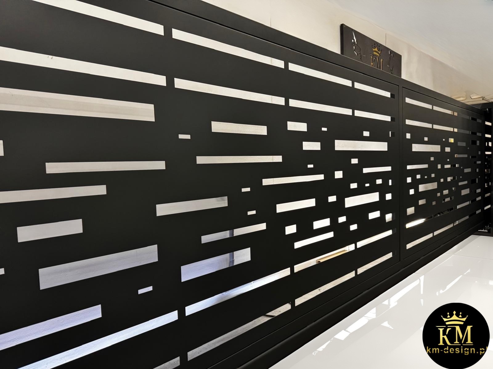 Czarna, przesuwna brama wjazdowa z nowoczesnym, geometrycznym wzorem wyciętym laserowo, widziana pod kątem. Błyszcząca, biała podłoga odbija światło.