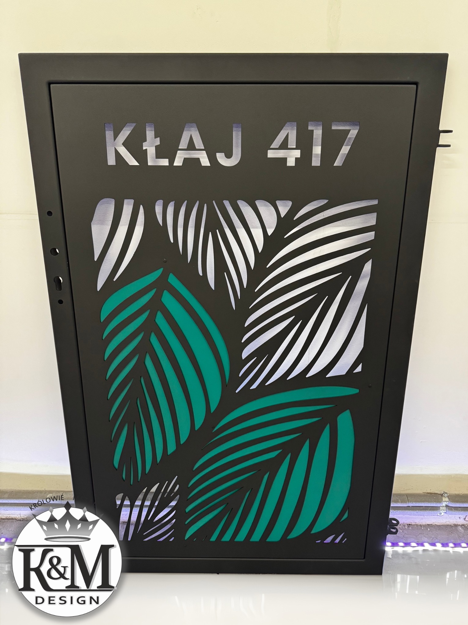Czarna metalowa skrzynka na listy z wycinanym laserowo wzorem liści palmowych i numerem domu 'KŁAJ 417', oświetlona od wewnątrz.