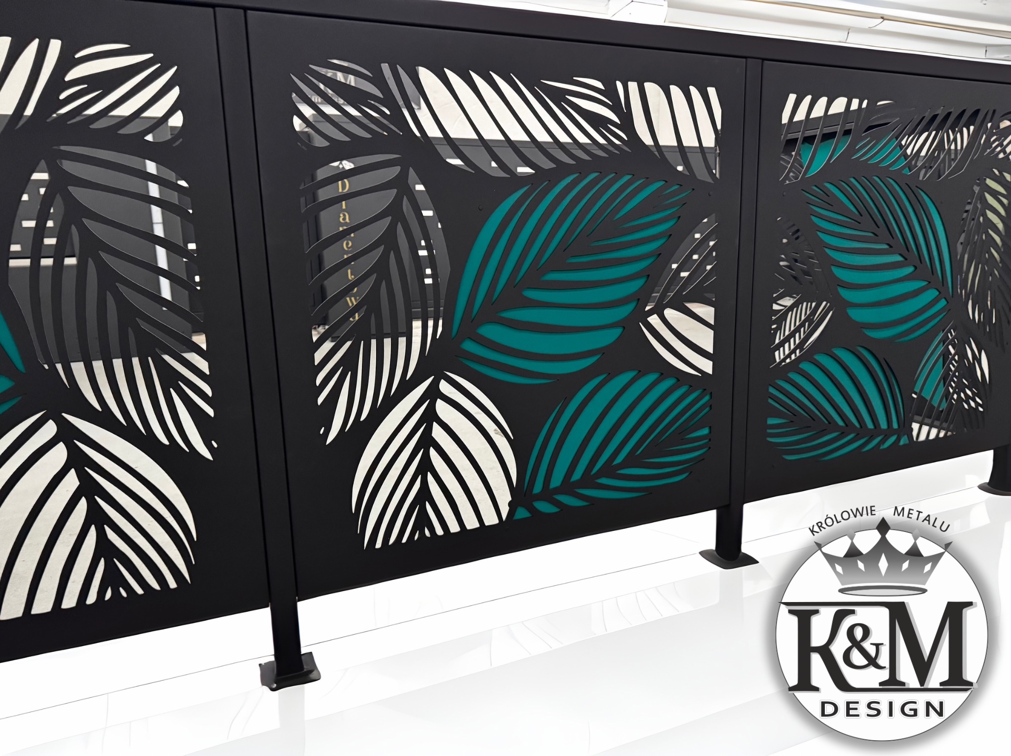 Czarna, metalowa balustrada z wycinanym wzorem liści palmowych, niektóre z nich podświetlone na turkusowo. Widoczny fragment podłogi i logo firmy.