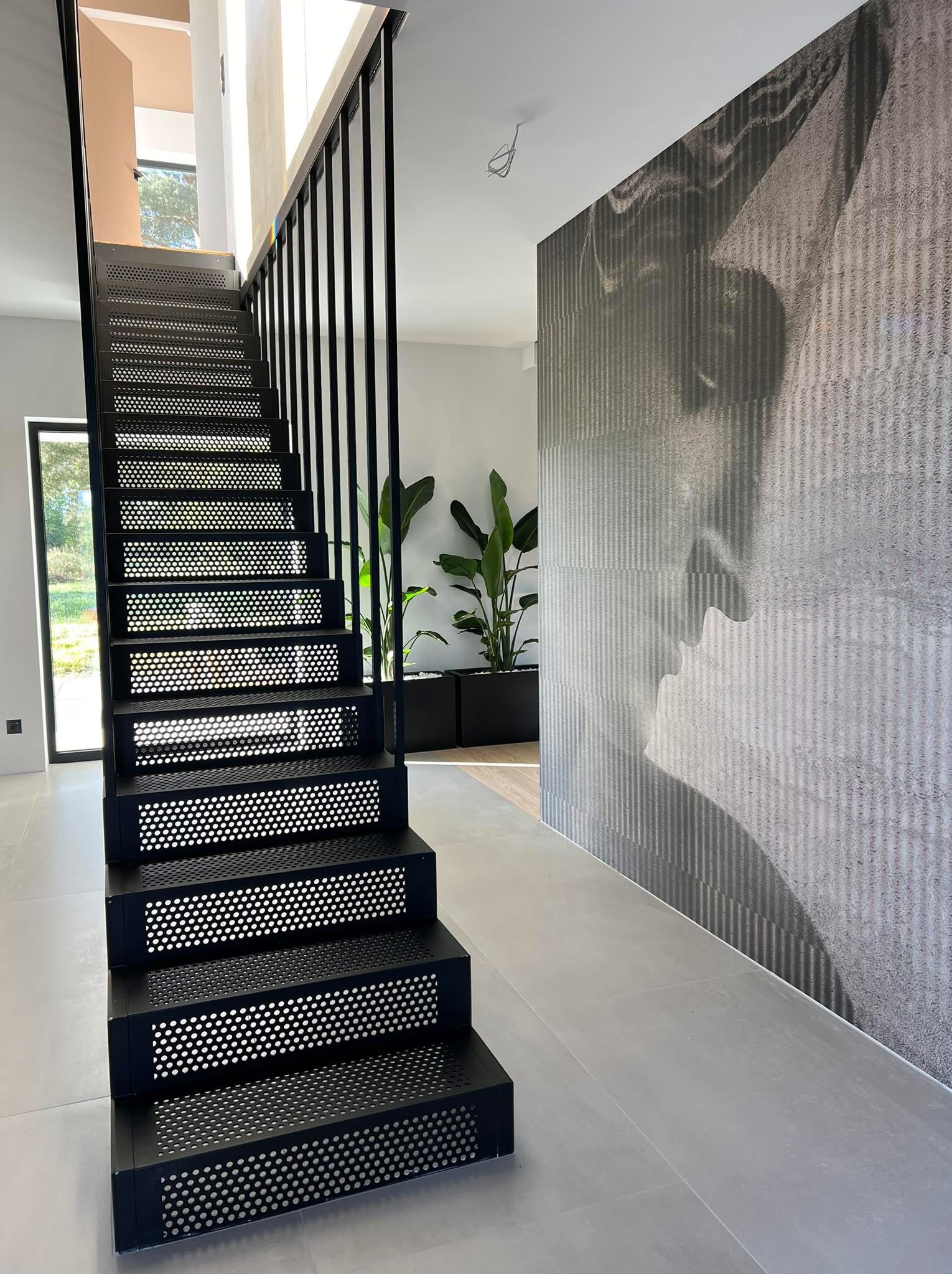 Nowoczesne schody metalowe w kolorze czarnym z perforowanymi stopniami, minimalistyczna balustrada, w tle ściana z abstrakcyjnym wzorem i rośliny doniczkowe.