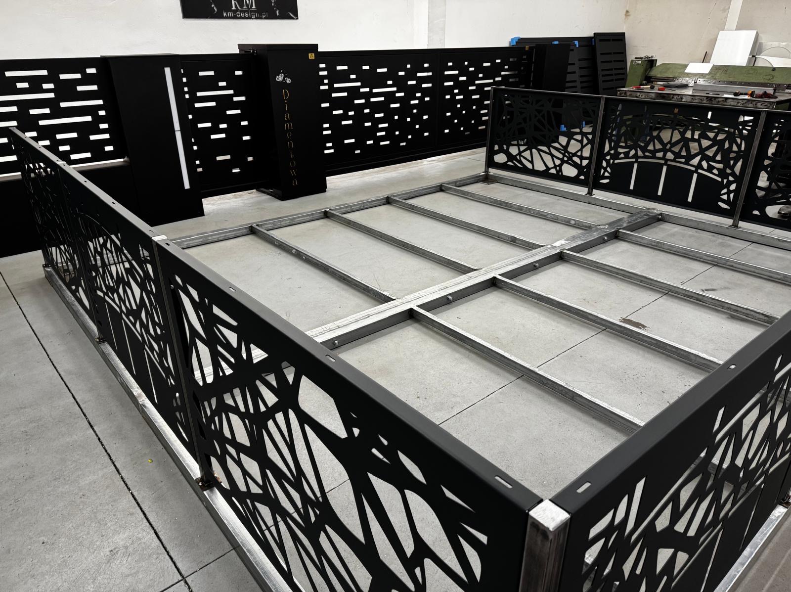Częściowo zmontowane, czarne, metalowe ogrodzenie z laserowo ciętym wzorem, oparte na metalowej ramie. W tle widoczne elementy innych ogrodzeń.