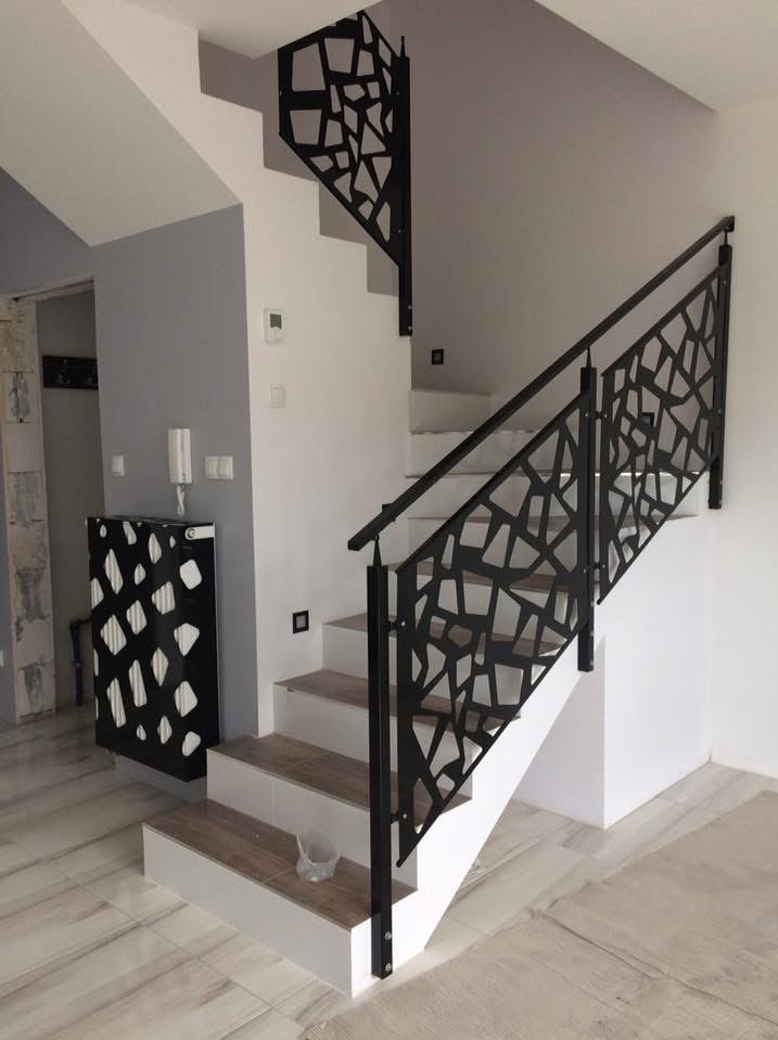 Nowoczesne schody z czarną, metalową balustradą o geometrycznym wzorze w jasnym wnętrzu. Widoczne płytki imitujące drewno na stopniach i podłodze.