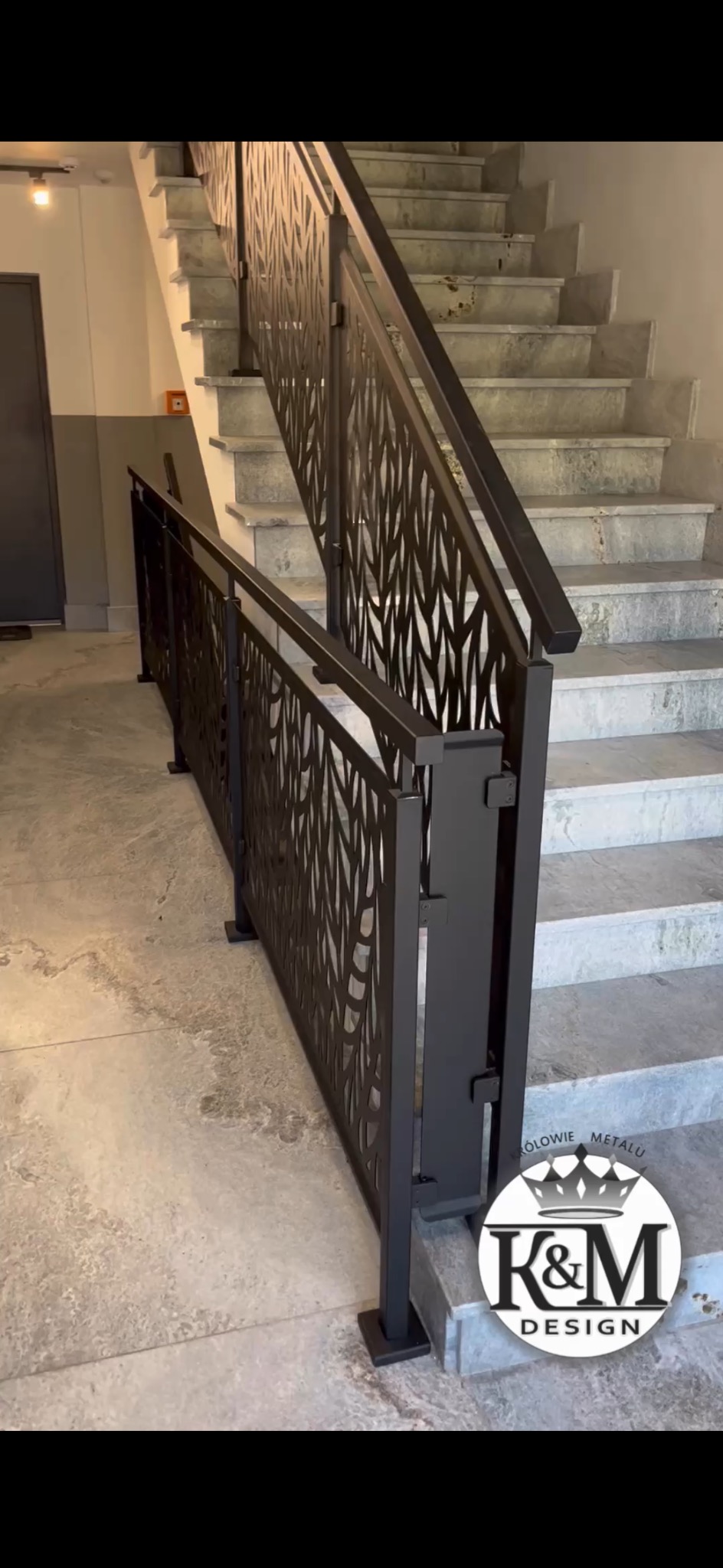 Nowoczesna, czarna balustrada metalowa schodowa z ażurowym wzorem liści, zamontowana wewnątrz budynku. Widoczne marmurowe schody i logo firmy.