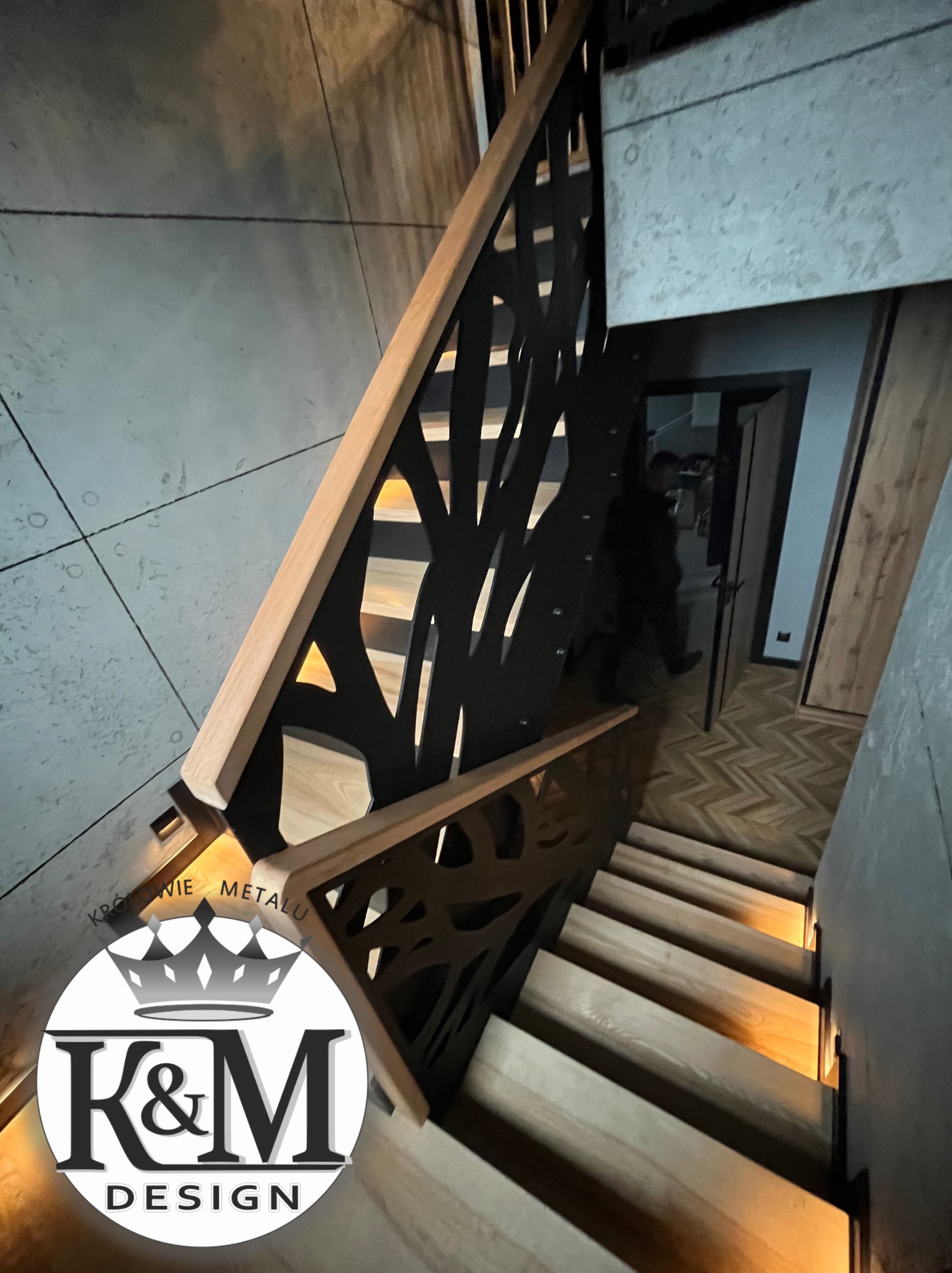 Nowoczesne schody z metalową balustradą o ażurowym wzorze, drewniane stopnie podświetlone od spodu. Widok z góry, w tle widoczny fragment wnętrza.