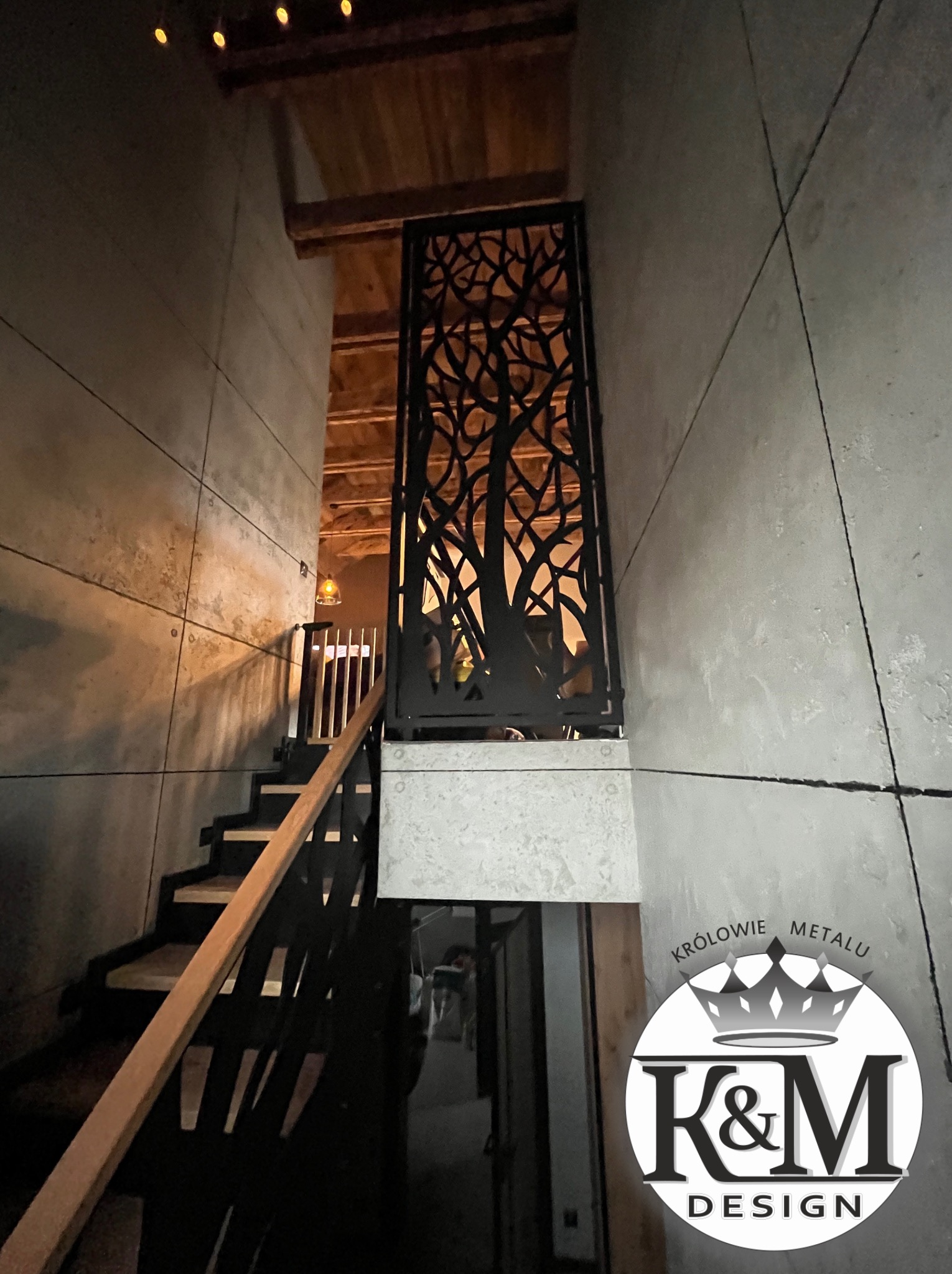 Metalowa balustrada schodowa z motywem drzewa, widoczna z dołu, w nowoczesnym wnętrzu z betonowymi ścianami i drewnianym sufitem. Unikalny wzór wycinany laserowo.
