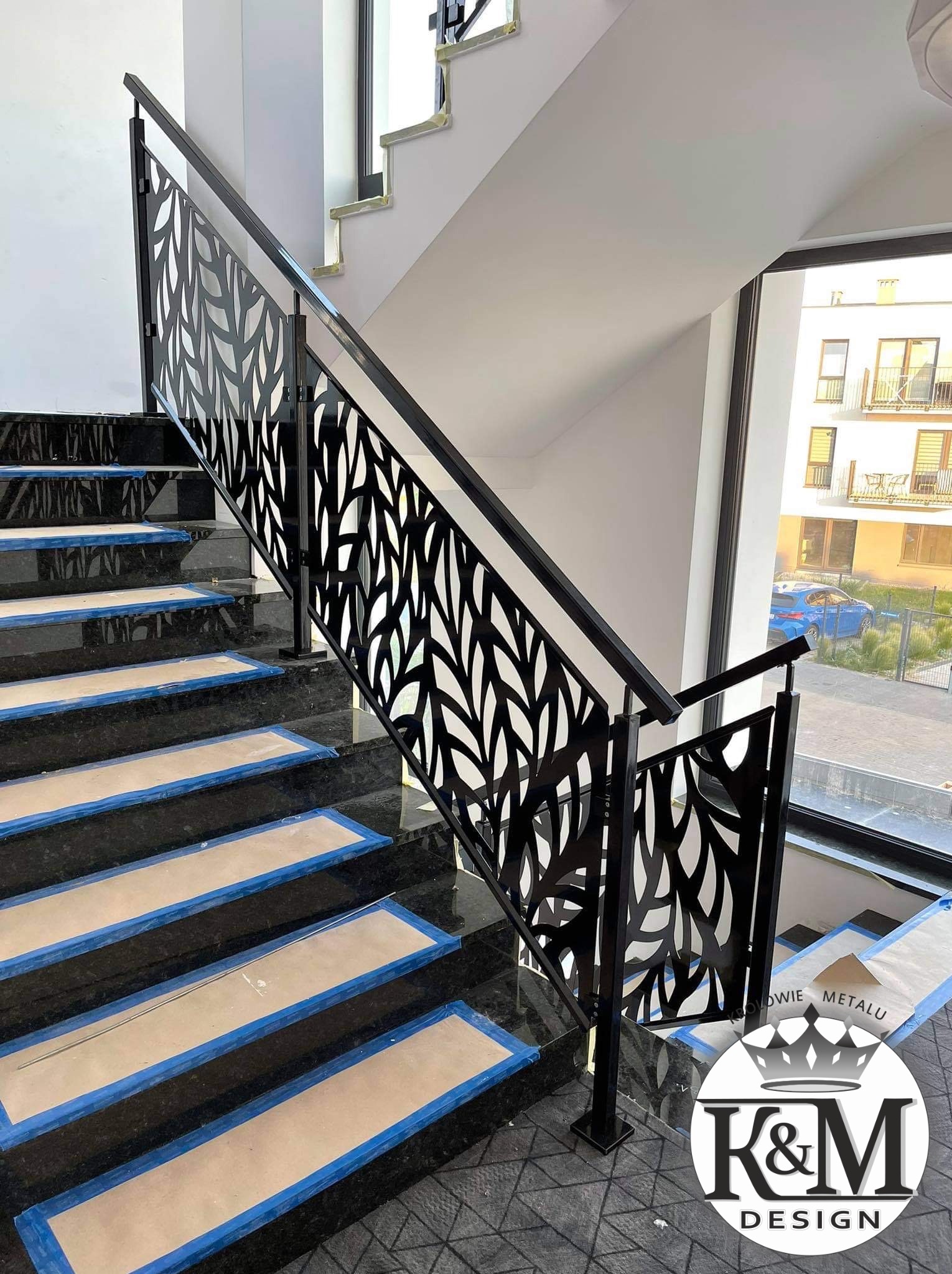 Nowoczesna, czarna balustrada metalowa z motywem liści na marmurowych schodach wewnątrz budynku, widok z dołu. Na stopniach zabezpieczenia.