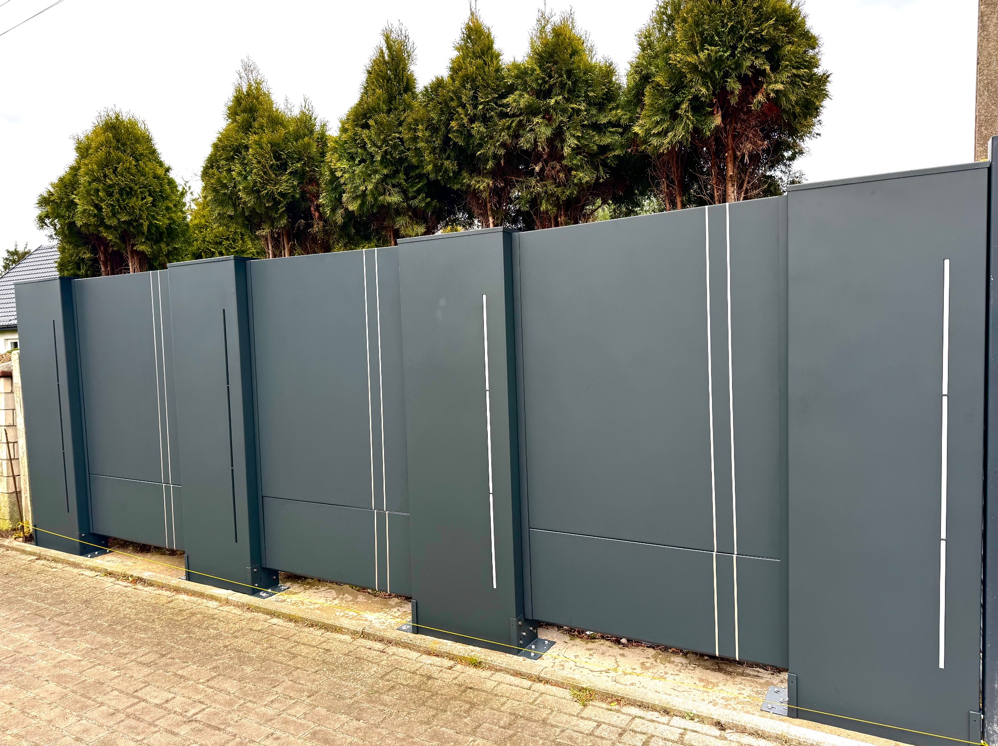 Nowoczesna, przesuwna brama wjazdowa w kolorze grafitowym z pionowymi, białymi zdobieniami, na tle zielonych tuj i kostki brukowej. Widoczne mocowania bramy.