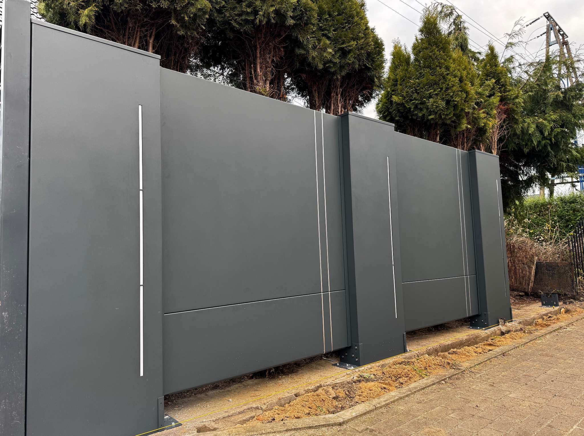 Nowoczesna, przesuwna brama wjazdowa w kolorze grafitowym z minimalistycznymi, pionowymi zdobieniami, widoczna z boku, na tle zielonych drzew i kostki brukowej.