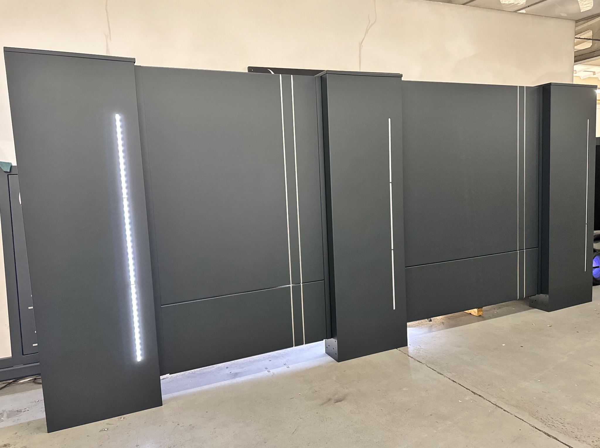 Nowoczesna, grafitowa brama wjazdowa z subtelnym oświetleniem LED, widoczna w hali produkcyjnej. Minimalistyczny design z pionowymi akcentami.