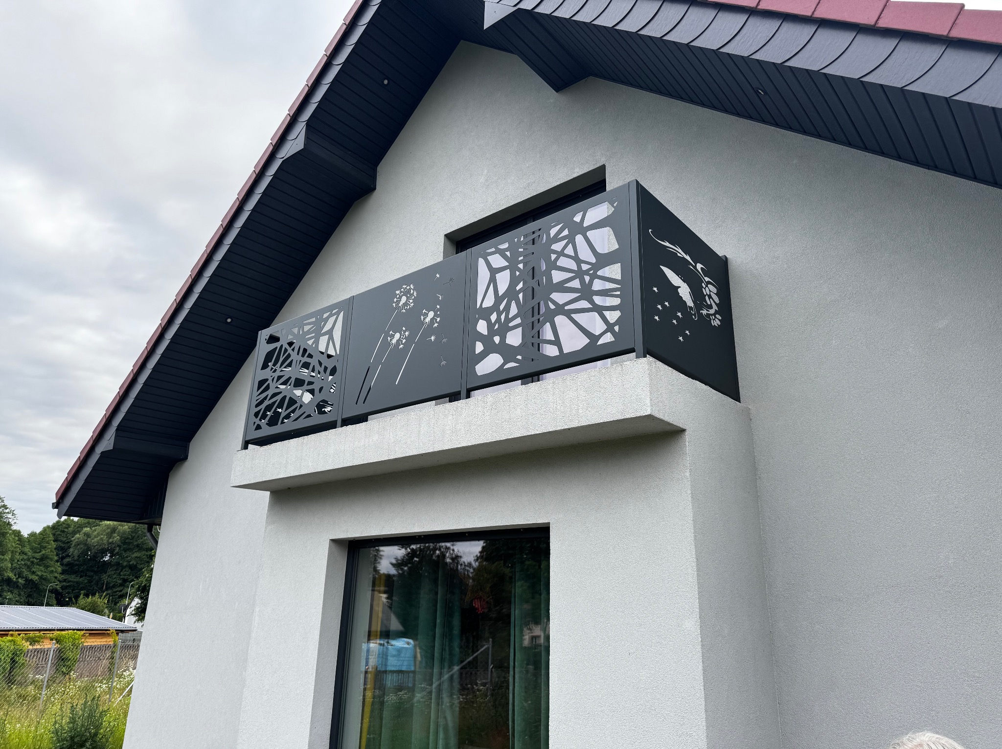 Nowoczesna, ażurowa balustrada balkonowa z motywem dmuchawca i motyla, zamontowana na balkonie domu z szarą elewacją i ciemnym dachem.