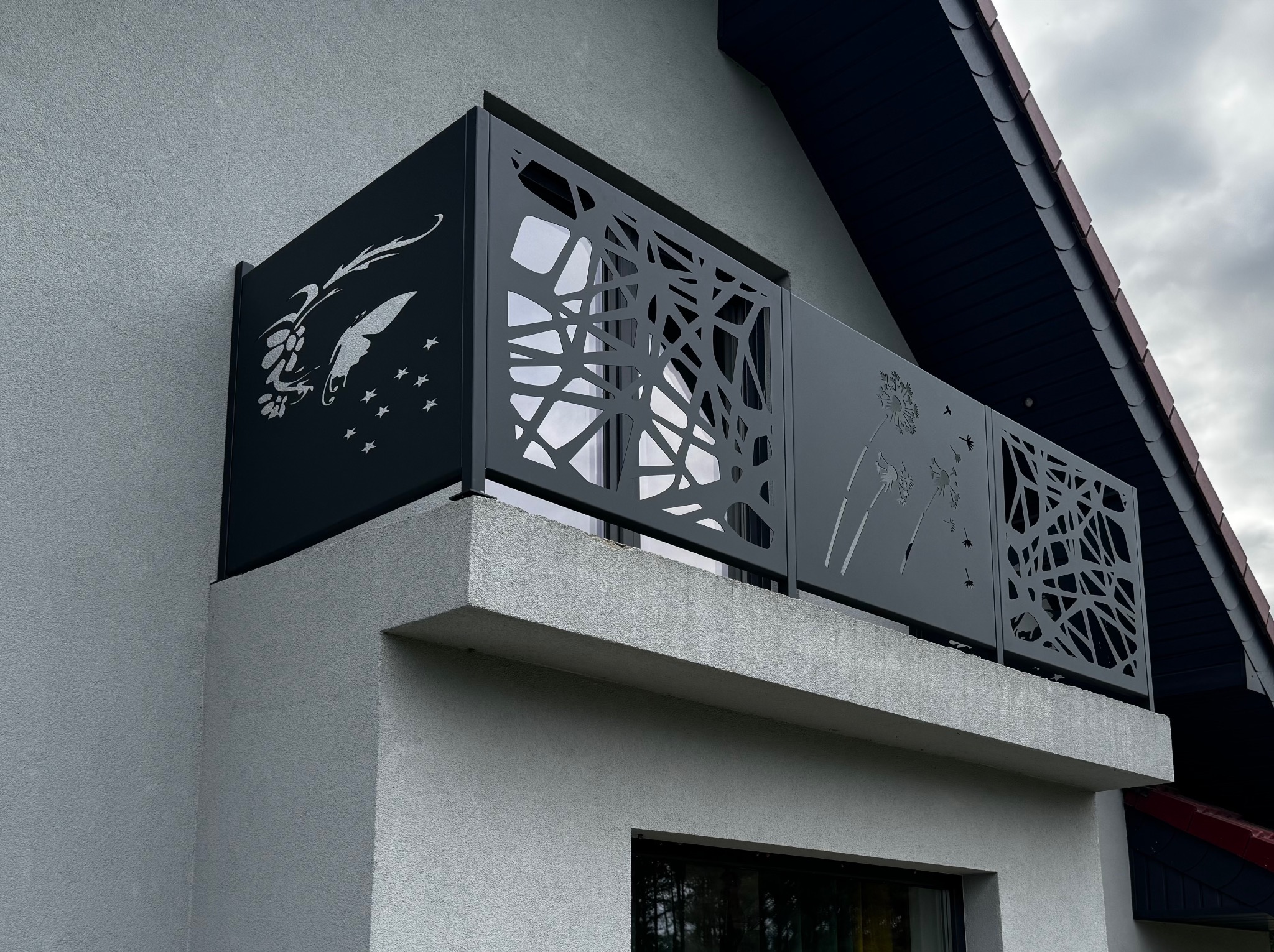 Czarna, ażurowa balustrada balkonowa z motywami roślinnymi i motyla, zamontowana na balkonie budynku z szarą elewacją. Nowoczesny design.