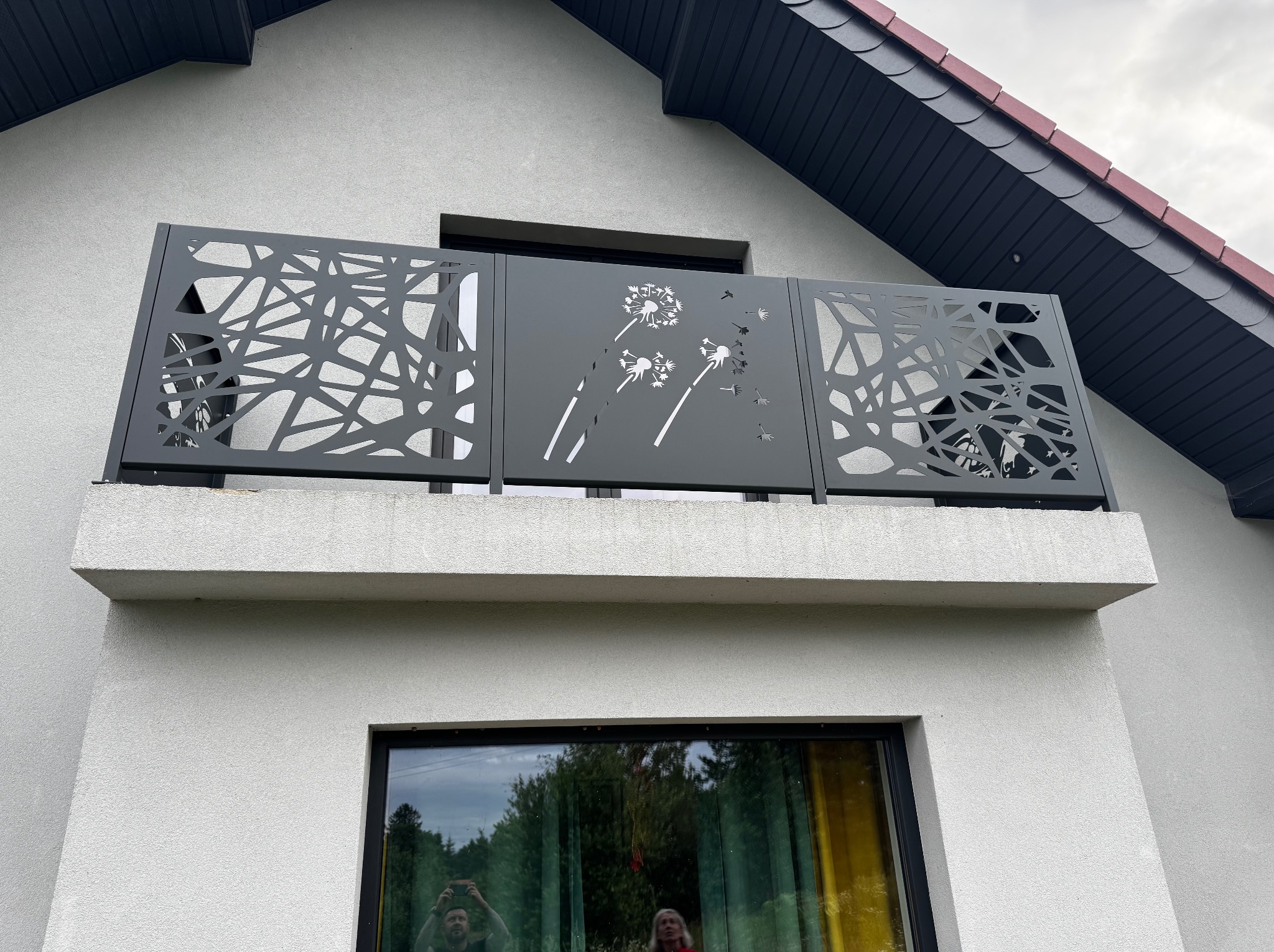 Nowoczesna, ażurowa balustrada balkonowa w kolorze grafitowym z motywem dmuchawców, zamontowana na balkonie domu z szarą elewacją. Widoczne odbicie osób w oknie.