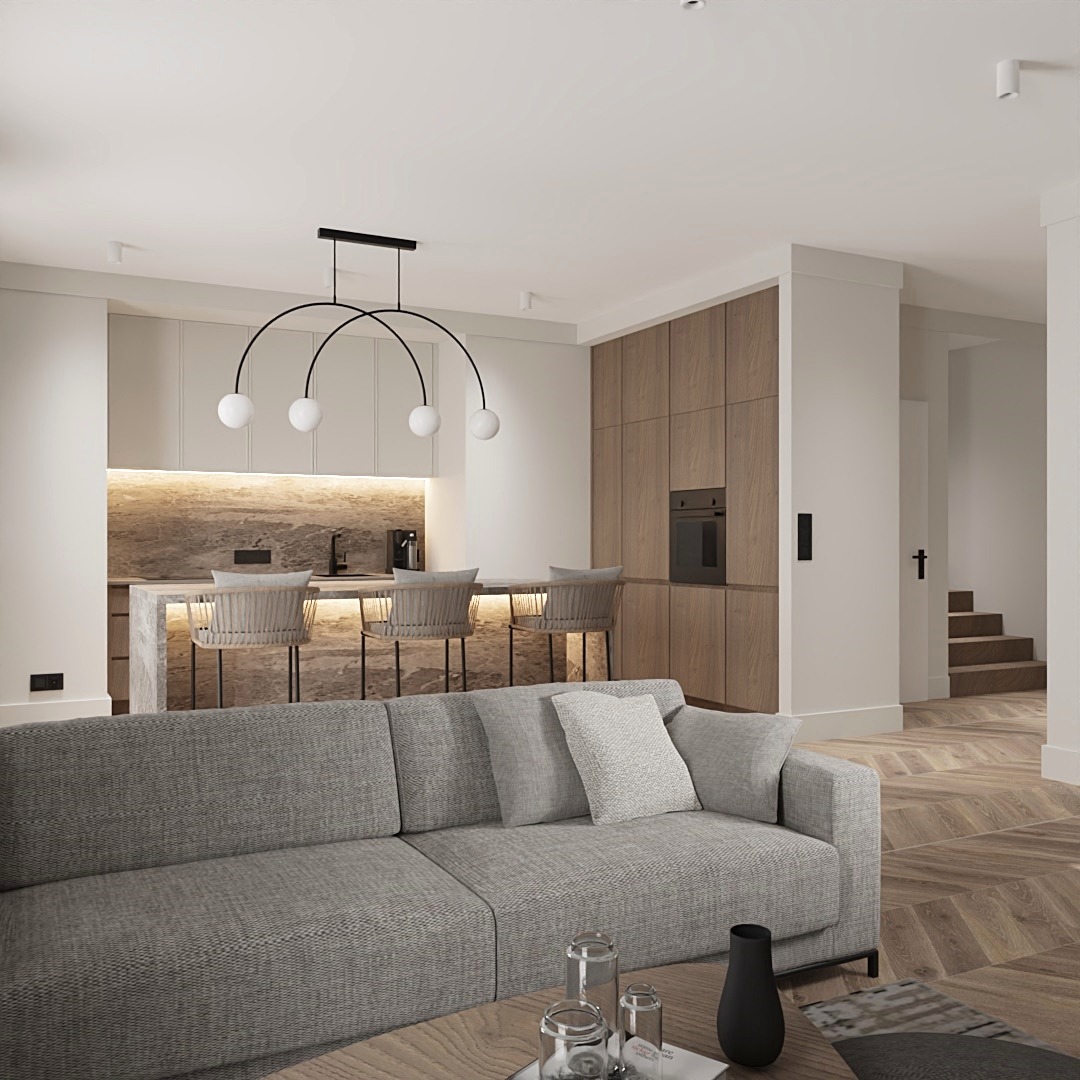 Nowoczesne wnętrze: szara sofa z poduszkami, okrągły stolik kawowy z dekoracjami, kuchnia z barkiem i krzesłami barowymi w tle, parkiet w jodełkę, minimalistyczna lampa wisząca z białymi kulami.