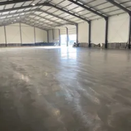 Świeżo wylana, jasnoszara posadzka w dużym, przemysłowym obiekcie w Raszkowie, z widocznymi śladami po maszynach i pracownikach w tle.